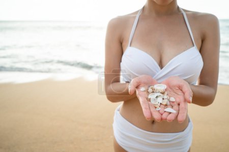 Foto de Woman in white bikini holding sea shell in hand on the beach. - Imagen libre de derechos