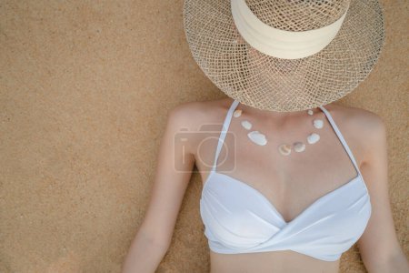 Téléchargez les photos : "Woman in white bikini lying on sand beach making necklace from sea shell, straw hat covering her face." - en image libre de droit