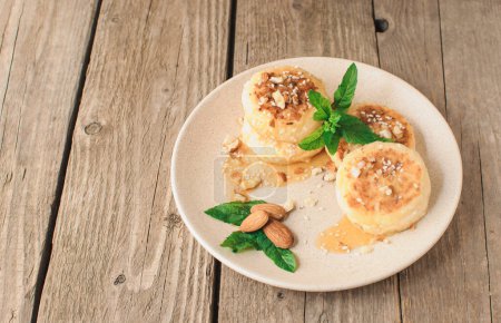 Téléchargez les photos : "Delicious breakfast - cottage cheese pancakes,cheesecakes, cottage cheese pancakes with almonds, mint and maple syrup in a beige plate.Useful dessert on a wooden table in rustic style.Selective focus." - en image libre de droit