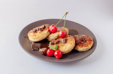 Foto de "Desayuno gourmet - tortitas de cuajada, pasteles de queso, tortitas de cuajada con cerezas y chocolate en un plato marrón. Postre saludable sobre un fondo blanco aislado" - Imagen libre de derechos