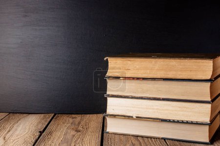 Photo for "books stacked on a wooden table in a rustic style on the background a school blackboard. The concept of welcome back to school. copy space" - Royalty Free Image