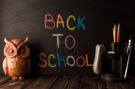 Photo for "Back to school, the concept of parenting. An assortment of consumables, owl, notebook, pencils, stand for pens. The inscription on the slate black chalk. School supplies on a wooden table, rustic." - Royalty Free Image