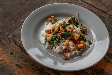 Téléchargez les photos : "Riz garni de basilic thaï frit sauté avec crevette naine servi dans un plat blanc sur une table en bois, nourriture de style thaï." - en image libre de droit