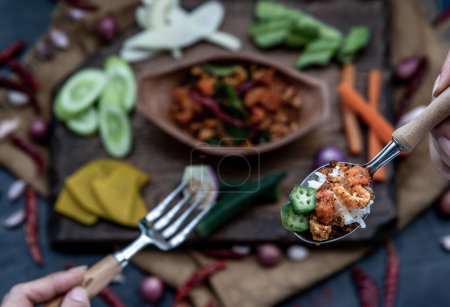 Photo for "The hand is using a spoon to scoop Pork Crackling Chili Paste with fried kaffir lime leaves with the ingredient and fresh vegetables on wood background." - Royalty Free Image