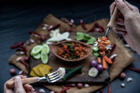 Photo for "The hand is using a spoon to scoop Pork Crackling Chili Paste with fried kaffir lime leaves with the ingredient and fresh vegetables on wood background." - Royalty Free Image