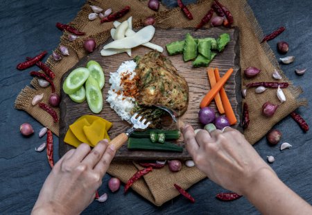 Téléchargez les photos : "Un style alimentaire thaïlandais local, La main utilise une cuillère pour ramasser l'omelette de pennata d'acacia frite ou les œufs cha-om sur du riz jasmin avec l'ingrédient et des légumes frais sur un fond en bois, " - en image libre de droit