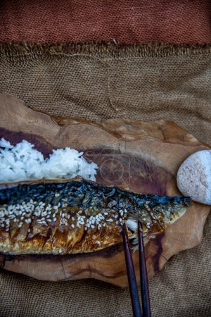 Téléchargez les photos : Main utilise des baguettes pour ramasser du poisson saba grillé sur du riz avec de la sauce teriyaki sur un fond de sac - en image libre de droit