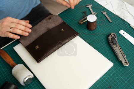 Téléchargez les photos : "Main des hommes travaillant sur le portefeuille en cuir dans son atelier. Processus de travail avec un cuir naturel marron. Artisan tenant un outillage artisanal." - en image libre de droit