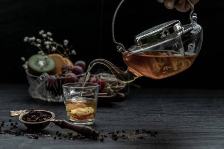 Téléchargez les photos : "Une belle composition avec du thé aux fruits chauds est versée de la théière en verre dans un verre avec une tisane et une feuille de thé." - en image libre de droit
