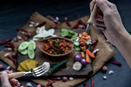 Photo for Hand using a spoon to scoop Pork Crackling Chili Paste with fried kaffir lime leaves with the ingredient and fresh vegetables on wood background - Royalty Free Image