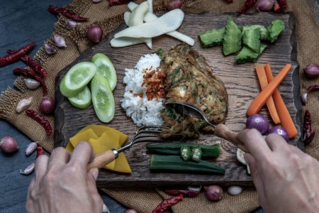 Foto de "A local Thai food style, The hand is using a spoon to scoop fried acacia pennata omelet or cha-om eggs on jasmine rice with the ingredient and fresh vegetables on a wooden background, " - Imagen libre de derechos