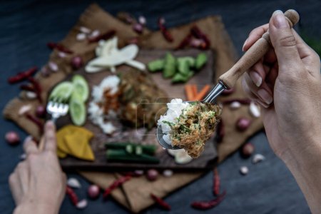 Photo for "A local Thai food style, The hand is using a spoon to scoop fried acacia pennata omelet or cha-om eggs on jasmine rice with the ingredient and fresh vegetables on a wooden background, " - Royalty Free Image