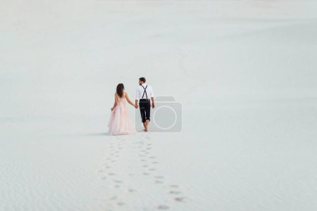 Foto de "pareja joven un chico en pantalones negros y una chica en un vestido rosa están caminando a lo largo de la arena blanca" - Imagen libre de derechos