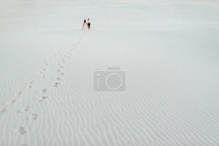 Foto de "pareja joven un chico en pantalones negros y una chica en un vestido rosa están caminando a lo largo de la arena blanca" - Imagen libre de derechos