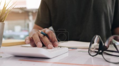 Téléchargez les photos : "Gros plan d'un homme d'affaires ou d'un comptable tenant un stylo à main travaillant sur une calculatrice pour calculer les données de l'entreprise, un document comptable et un ordinateur portable au bureau, concept d'entreprise" - en image libre de droit