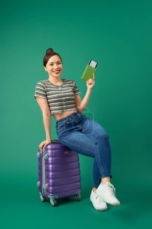 Foto de Jovencita alegre asiática sentada en la maleta y con pasaporte, billete de avión para viajar sobre fondo verde
. - Imagen libre de derechos