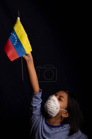 Foto de Retrato de una niña venezolana usando máscara médica y sosteniendo con suerte la bandera de Venezuela - Imagen libre de derechos