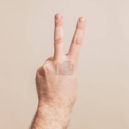 Téléchargez les photos : Main masculine montrant le signe de victoire sur fond beige - en image libre de droit