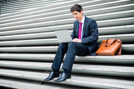 Photo for "Young businessman using a laptop while sitting down outdoors " - Royalty Free Image
