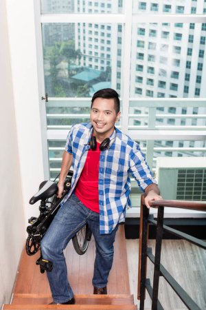 Foto de "joven asiático hombre llevar plegable bicicleta arriba la escaleras a apartamento" - Imagen libre de derechos