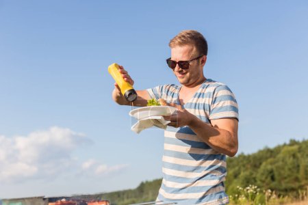 Téléchargez les photos : "Jeune homme ajoutant de la moutarde à son assiette de barbecue" - en image libre de droit