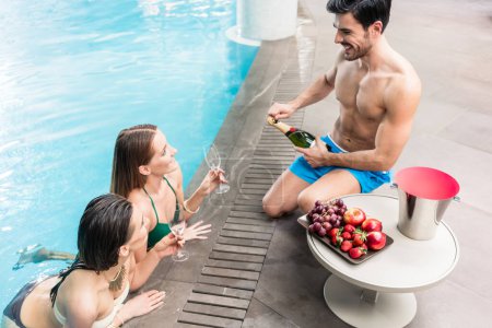 Téléchargez les photos : "Jeune homme servant avec champagne deux femmes à la piscine d'un hôtel de luxe" - en image libre de droit