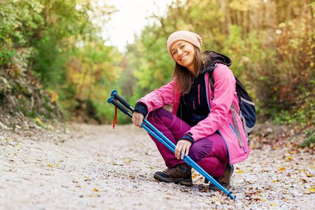 Photo for "Hiker girl crouching on a wide trail in the mountains. Backpacker with pink jacket in a forest. Healthy fitness lifestyle outdoors." - Royalty Free Image