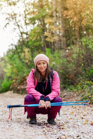 Photo for "Hiker girl crouching on a wide trail in the mountains. Backpacker with pink jacket in a forest. Healthy fitness lifestyle outdoors." - Royalty Free Image