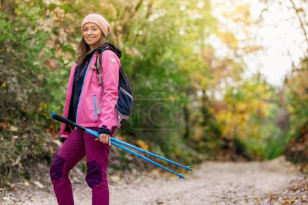 Photo for "Hiker girl standing on a wide trail in the mountains. Backpacker with pink jacket in a forest. Healthy fitness lifestyle outdoors." - Royalty Free Image