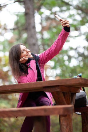 Téléchargez les photos : "Une randonneuse reposant sur un banc dans la forêt. Sac à dos avec veste rose prenant selfie avec smartphone." - en image libre de droit
