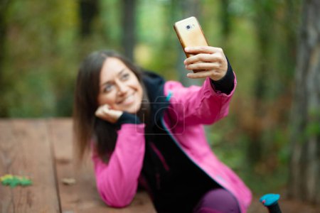 Téléchargez les photos : "Une randonneuse reposant sur un banc dans la forêt. Sac à dos avec veste rose prenant selfie avec smartphone." - en image libre de droit