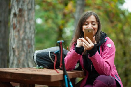 Téléchargez les photos : "Une randonneuse reposant sur un banc dans la forêt. Sac à dos avec veste rose tenant le téléphone portable." - en image libre de droit