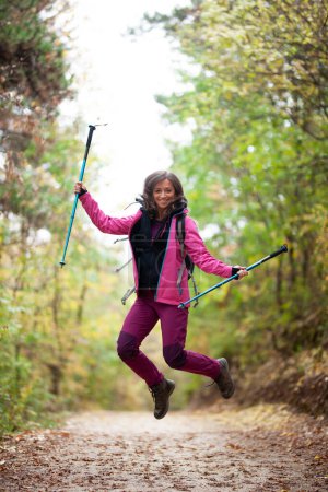 Photo for "Hiker girl jumping on a trail in the mountains. Backpacker with hiking poles and pink jacket in a forest. Happy lifestyle outdoors." - Royalty Free Image