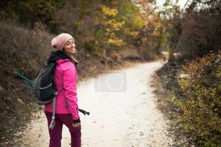 Téléchargez les photos : "Randonneuse pédestre marchant sur un sentier dans les montagnes. Vue arrière du sac à dos avec veste rose dans une forêt. Style de vie sain en plein air." - en image libre de droit