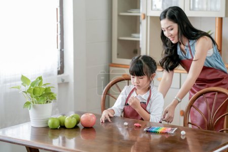 Photo for "Little girl enjoy to play with play dough in kitchen on the table while her mother try to dress her with apron. Asian family with happiness concept." - Royalty Free Image