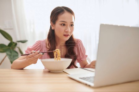Téléchargez les photos : Portrait de jeune fille asiatique utilisant un ordinateur portable tout en mangeant des nouilles à la maison - en image libre de droit
