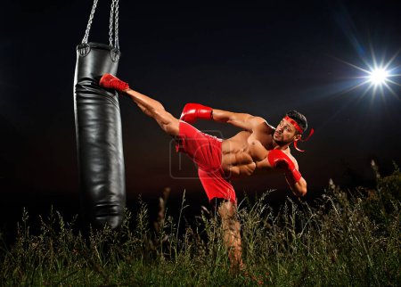 Photo for Sportsman is training on a black night background, boxer in red shorts and red boxing gloves, muscles, muscular body, night training, training in open space,side kick to a punching bag, black punching bag. - Royalty Free Image