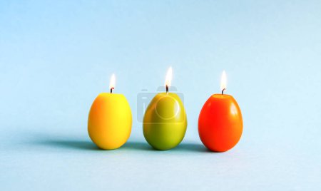 Foto de Bright burning paraffin candles in the shape of colorful Easter eggs on blue background - Imagen libre de derechos