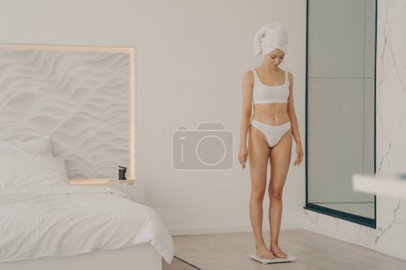 Photo for "Attractive slim young girl standing on scales in bedroom" - Royalty Free Image