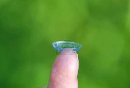 Téléchargez les photos : "Lentille de contact transparente sur le bout des doigts sur fond vert flou été nature" - en image libre de droit