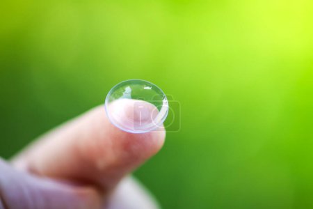 Téléchargez les photos : Lentille de contact transparente sur le bout des doigts sur fond vert flou été nature - en image libre de droit