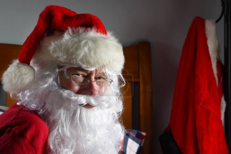 Photo for Santa Claus sitting on bed with his Red Suit hanging from a hook in the background - Royalty Free Image