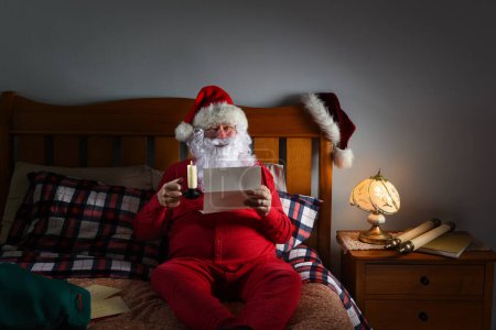 Photo for Santa Claus in his red long johns reading letters by candlelight on top of his bed at the North Pole - Royalty Free Image