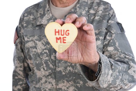 Foto de Primer plano de un soldado sosteniendo una galleta de San Valentín en forma de corazón con las palabras Abrázame escrito en glaseado rojo. - Imagen libre de derechos