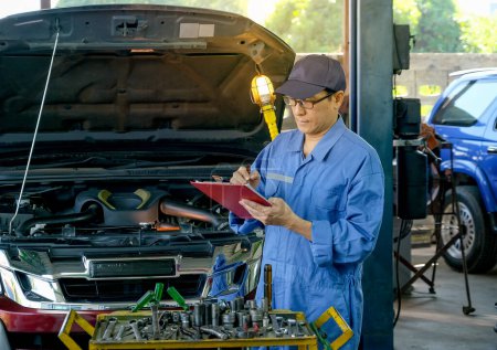 Foto de "Retrato de mecánico automotriz asiático con uniforme azul comprobar la lista a bordo para solucionar el problema del coche en el garaje." - Imagen libre de derechos
