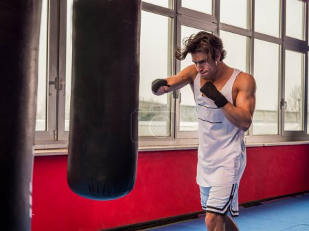 Foto de Entrenamiento de hombre deportivo muscular con saco de boxeo mientras boxea - Imagen libre de derechos
