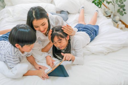 Téléchargez les photos : "Flou doux de mère asiatique et ses enfants s'amusent avec tablette dans la chambre." - en image libre de droit