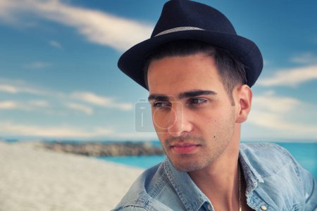 Téléchargez les photos : Beau jeune, homme en denim chemise et chapeau de fedora assis à la plage - en image libre de droit