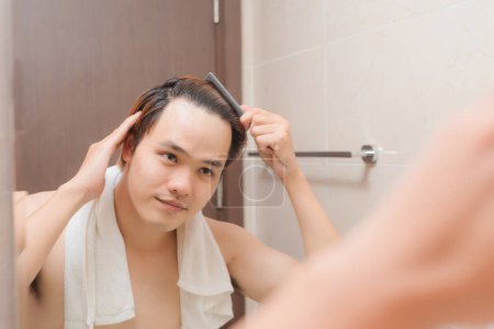 Téléchargez les photos : " sourire jeune homme regardant pour miroir et brosser les cheveux avec peigne à la maison salle de bain" - en image libre de droit