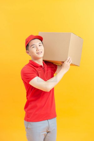 Foto de Joven hombre de entrega logística sonriente en uniforme rojo sosteniendo la caja en el fondo de color - Imagen libre de derechos